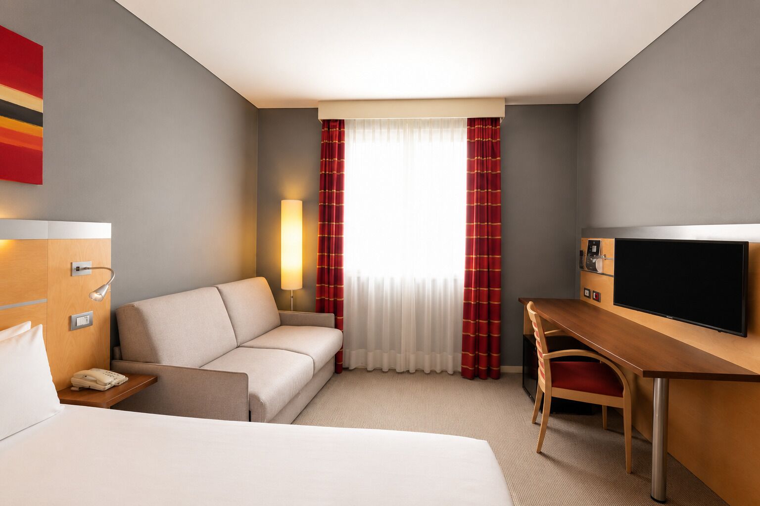 A hotel room with a bed, a sofa, a desk with a TV, a lamp, and a window with curtains.