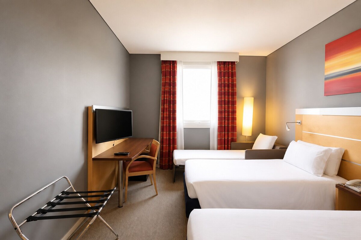 A hotel room with a desk, two beds, a TV, a lamp, and a window with curtains.