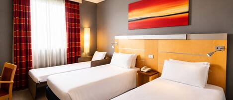A hotel room with two beds, a desk, a chair, a lamp, and a painting on the wall.