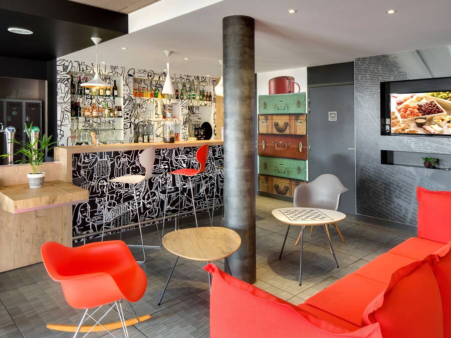 A modern bar area with a black and white graffiti wall, a wooden counter, and a variety of bottles and glasses. There are red and grey chairs, a small round table, and a television mounted on the wall displaying food.