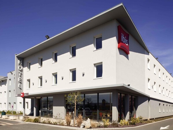 A modern hotel building with a flat roof, multiple windows, and a prominent red 'ibis' sign.