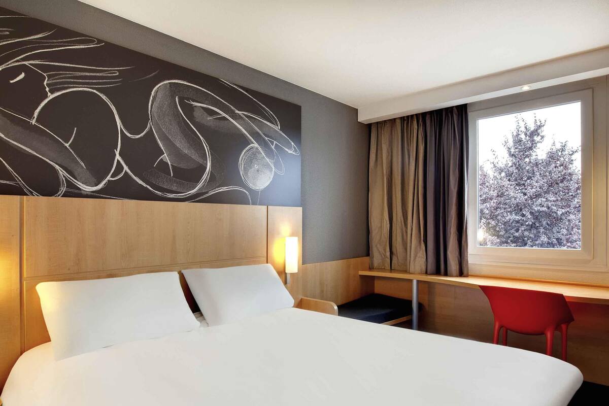 A hotel room with a large bed, a desk with a chair, a window with curtains, and a wall with a chalkboard drawing.