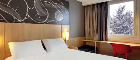 A hotel room with a large bed, a desk with a chair, a window with curtains, and a wall with a chalkboard drawing.