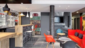 Un espace bar moderne avec un comptoir en bois, de nombreux tabourets et une variété de bouteilles. On y trouve des chaises rouges et bleues, un canapé rouge et une chaise noire avec un jeu d’échecs.