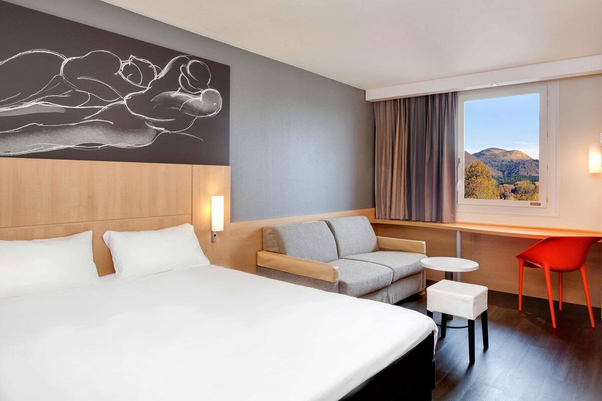 A hotel room with a bed, a sofa, a small table, and a chair. There is a window with a view of hills and trees.
