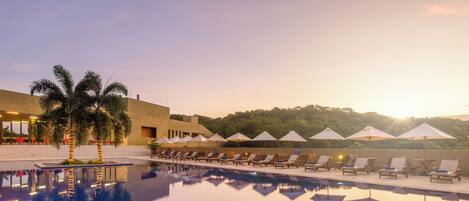 A pool with lounge chairs and umbrellas, palm trees, and a building with a flat roof.