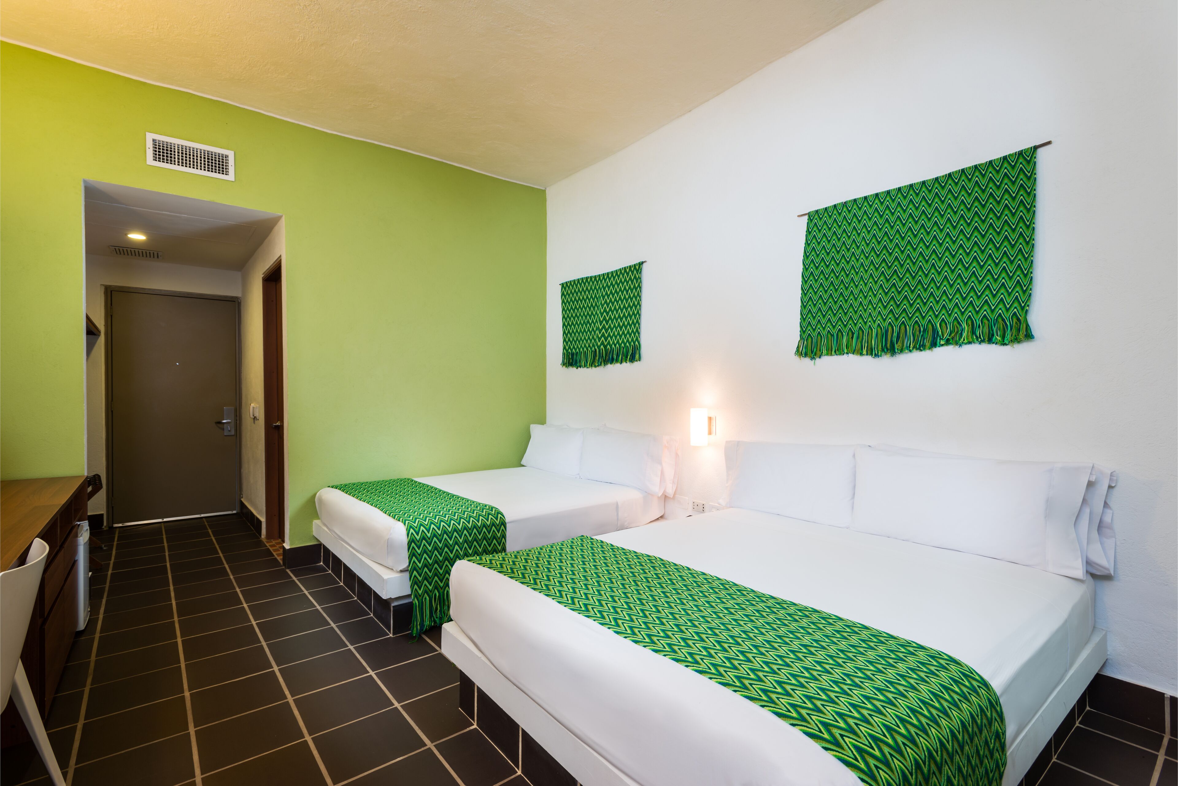 A hotel room with two beds, green and white bedding, a small desk, and a door leading to a bathroom.