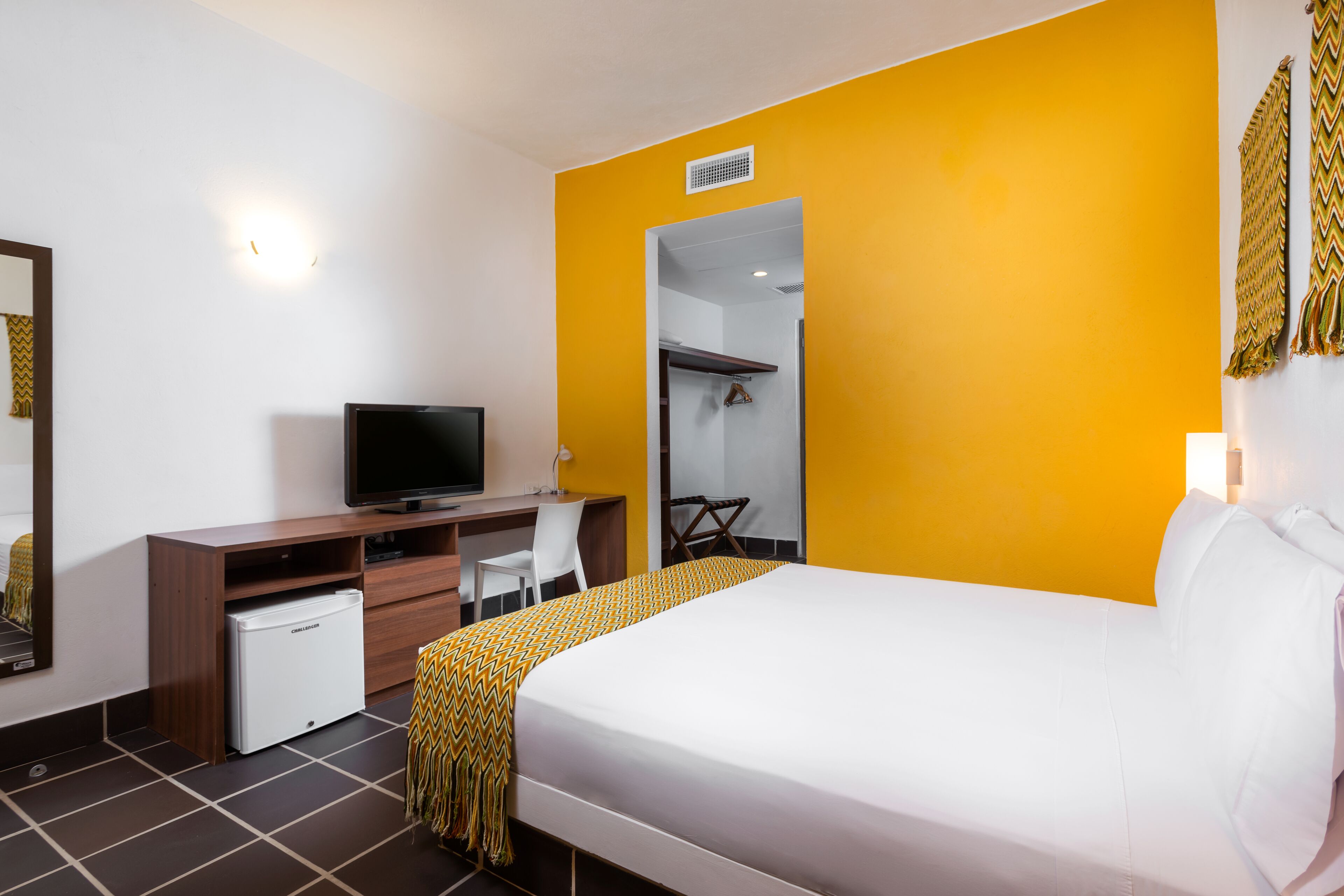 A hotel room with a bed, a desk with a television, a mini-fridge, and a wardrobe.