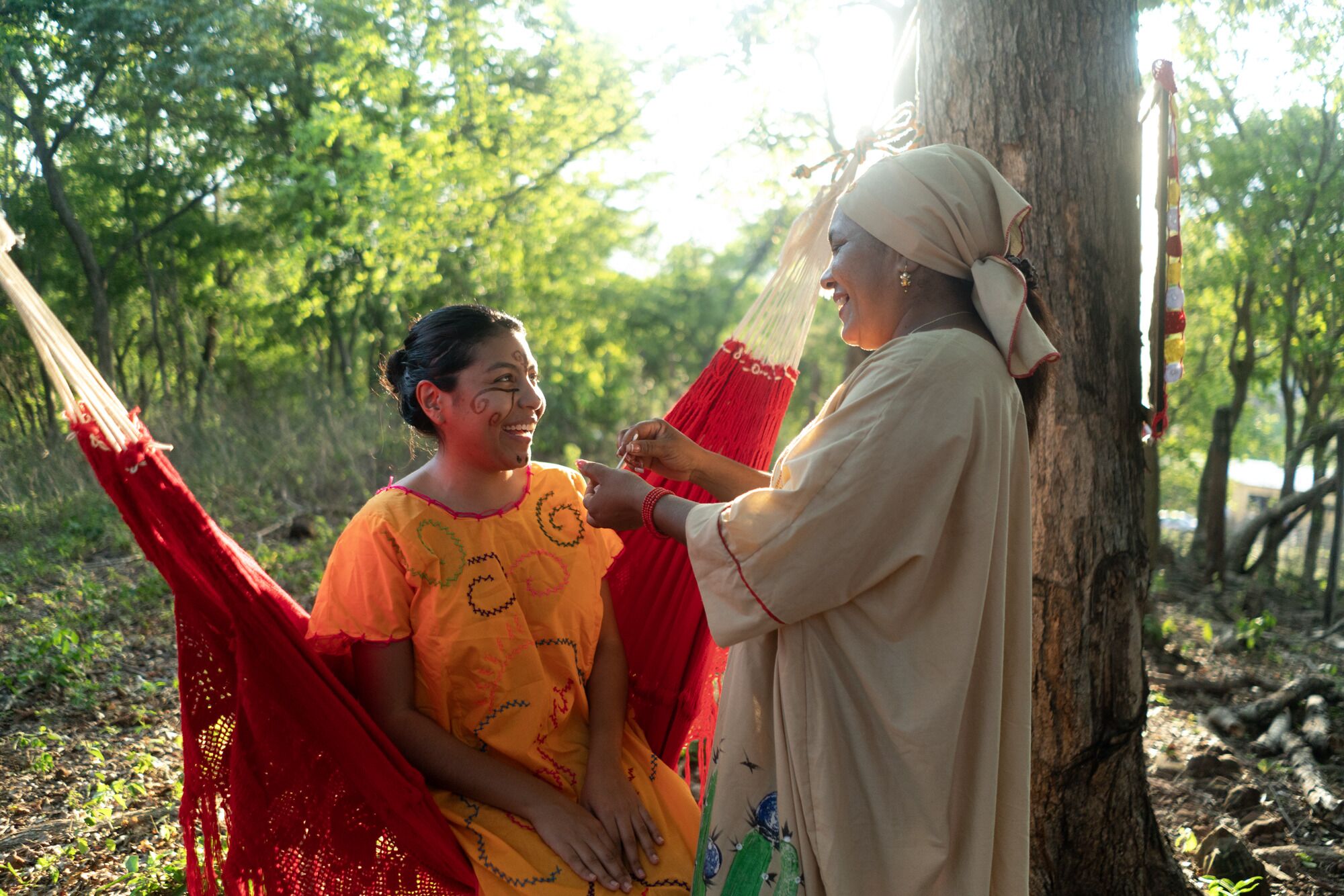 A person in a beige garment and another in a yellow garment sitting in a hammock outdoors.