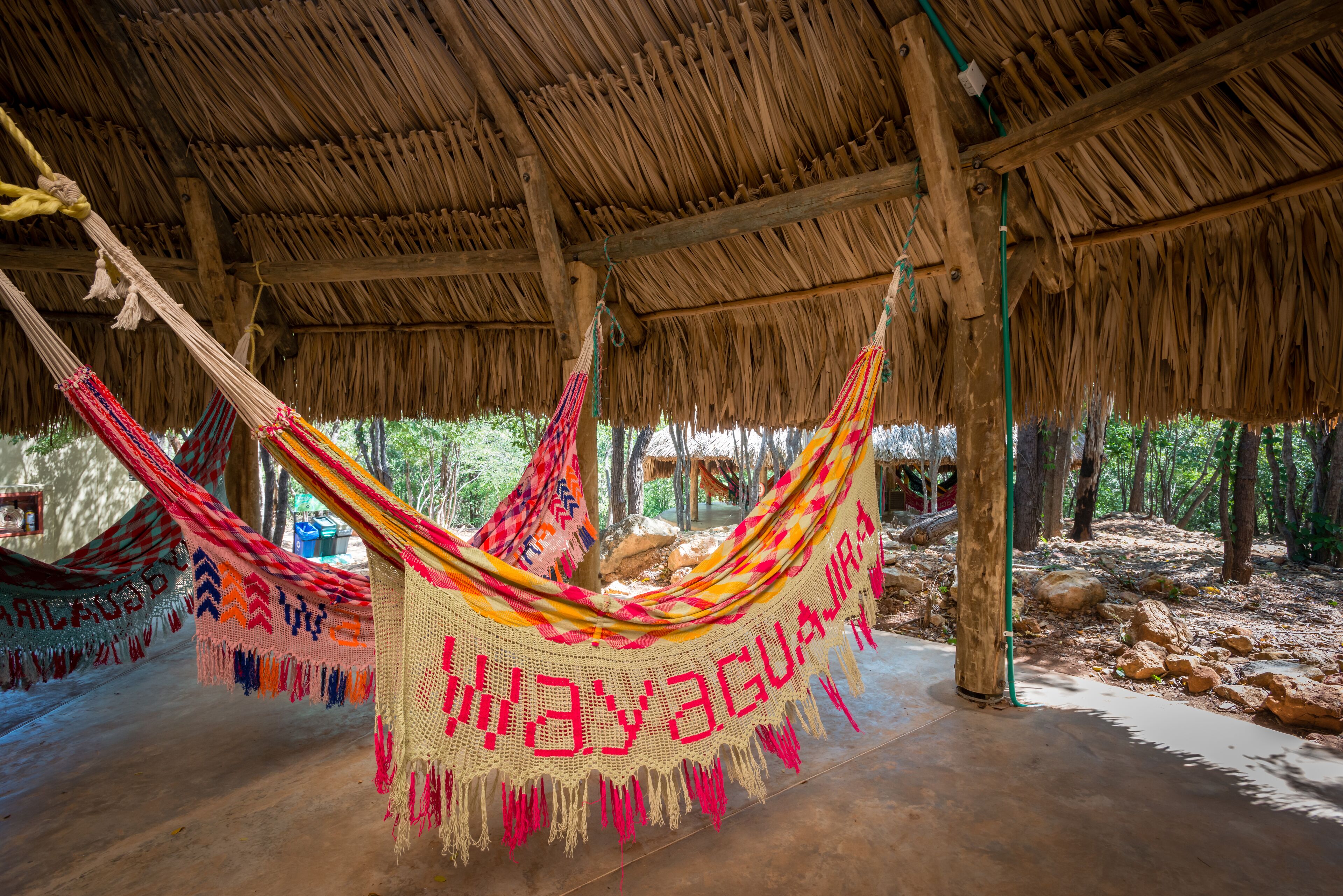 A hammock under a thatched roof structure with a woven mat hanging from the ceiling.