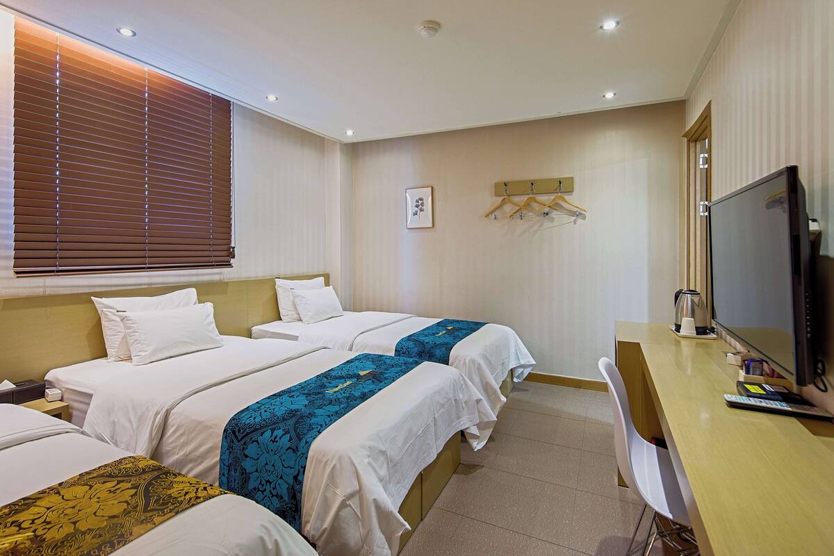 Habitación de hotel con dos camas, un escritorio, televisor y ventana con persianas.