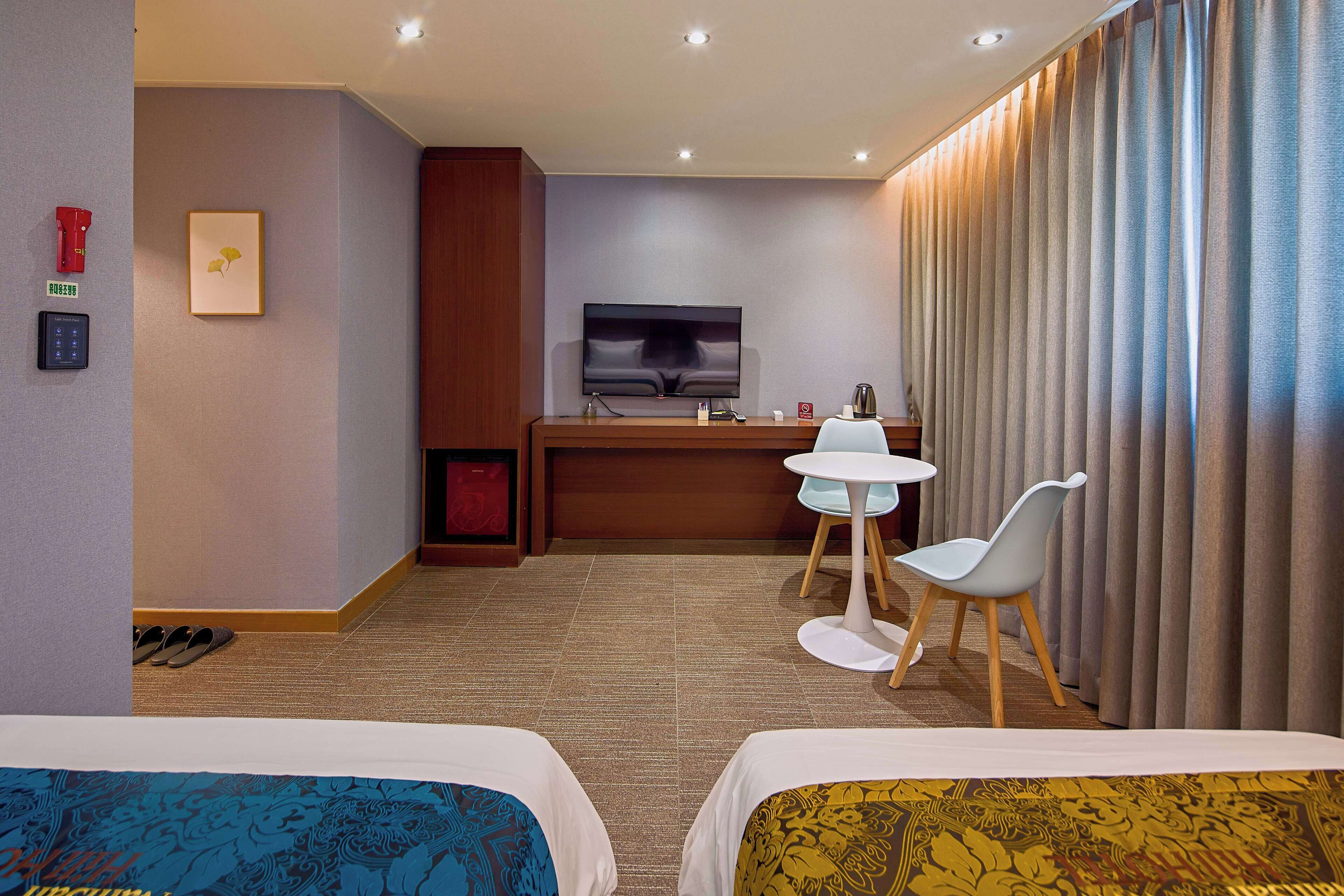 Une chambre d’hôtel avec deux lits, un bureau avec une télévision, une petite table avec une chaise et un rideau sur le côté droit.