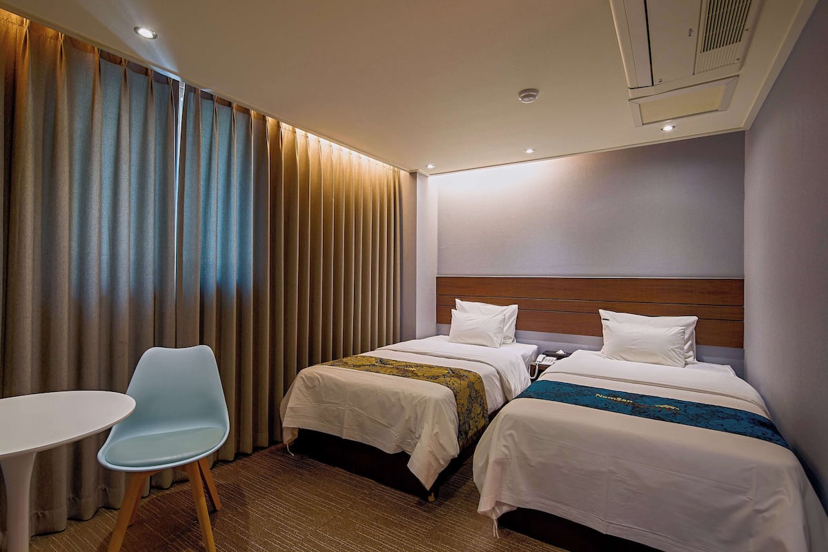 Habitación de hotel con dos camas, una mesita, una silla y un ventanal con cortinas.