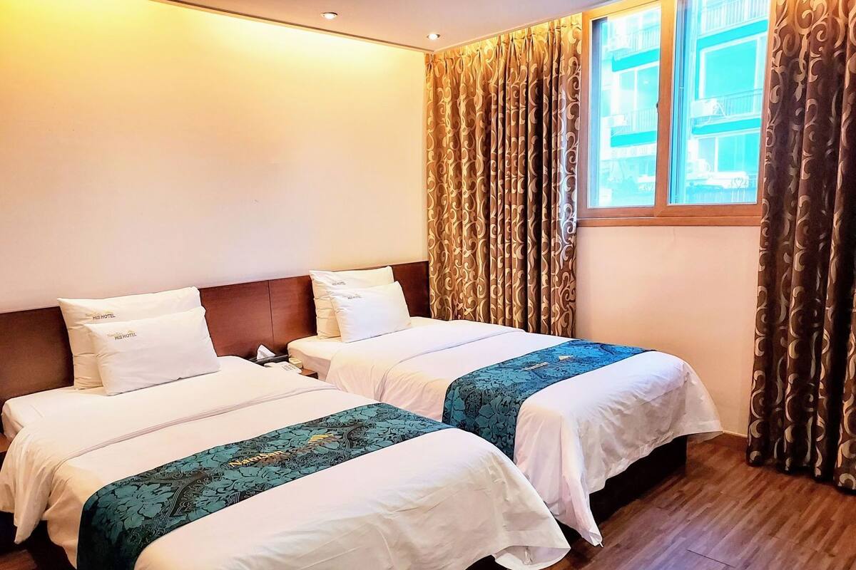 Habitación de hotel con dos camas, cabecera de madera, ventana con cortinas y piso de madera.