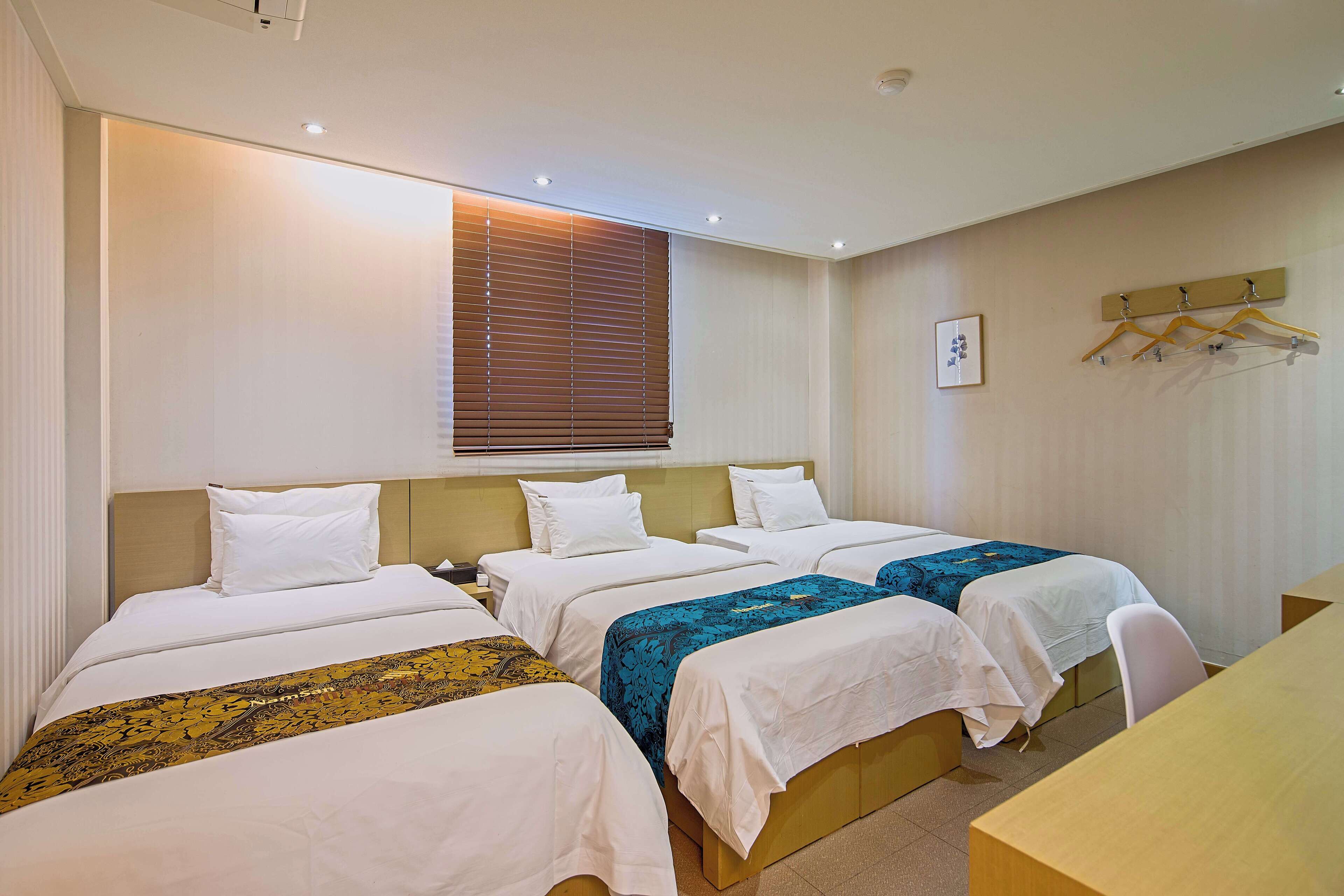 Habitación de hotel con tres camas individuales, cabecera de madera, mesita de noche, escritorio y perchero de pared.