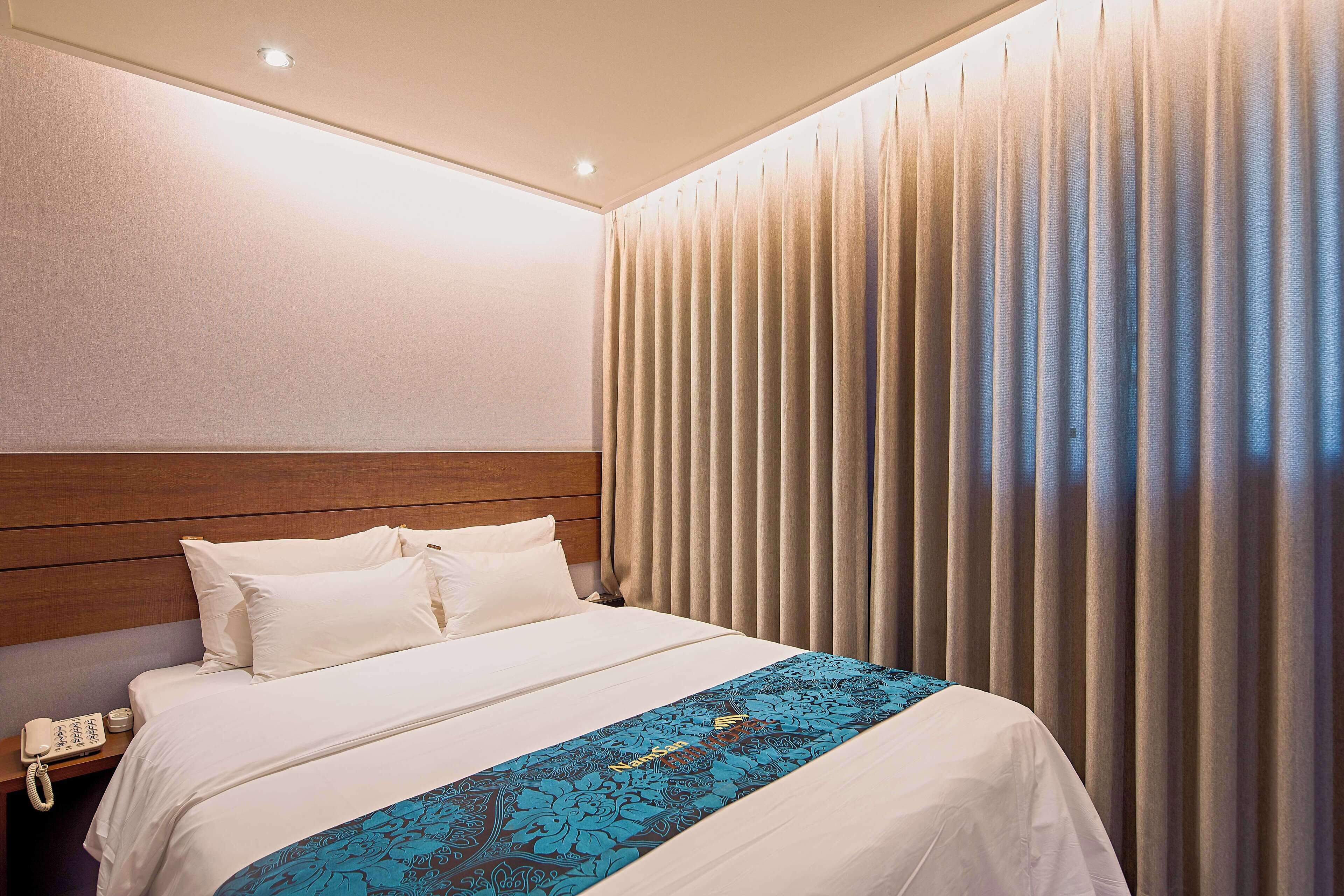 Quarto de hotel com uma cama bem arrumada, cabeceira de madeira, telefone na mesa de cabeceira e janela com persianas verticais.