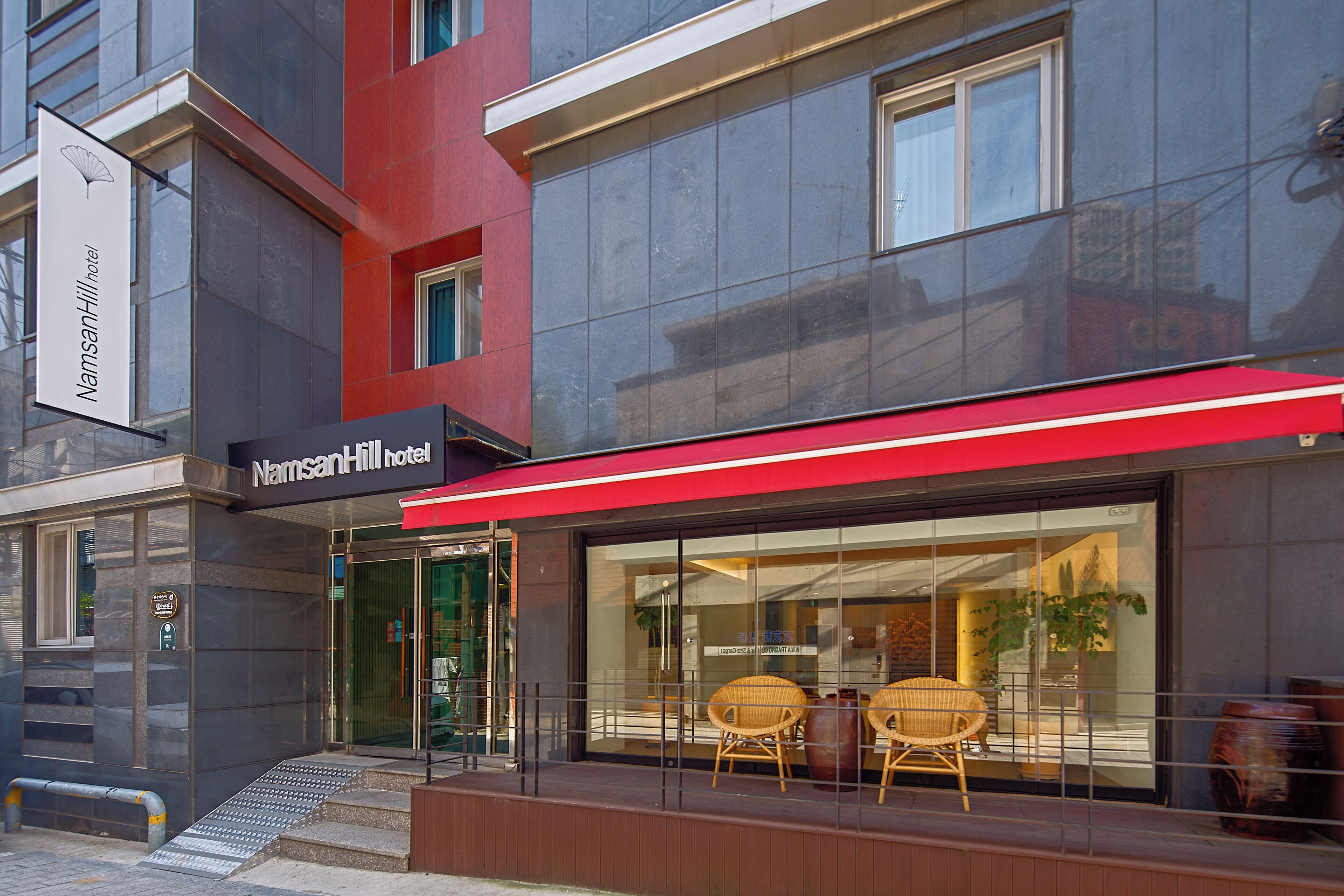 A hotel entrance with a red awning, glass doors, and wicker chairs outside.