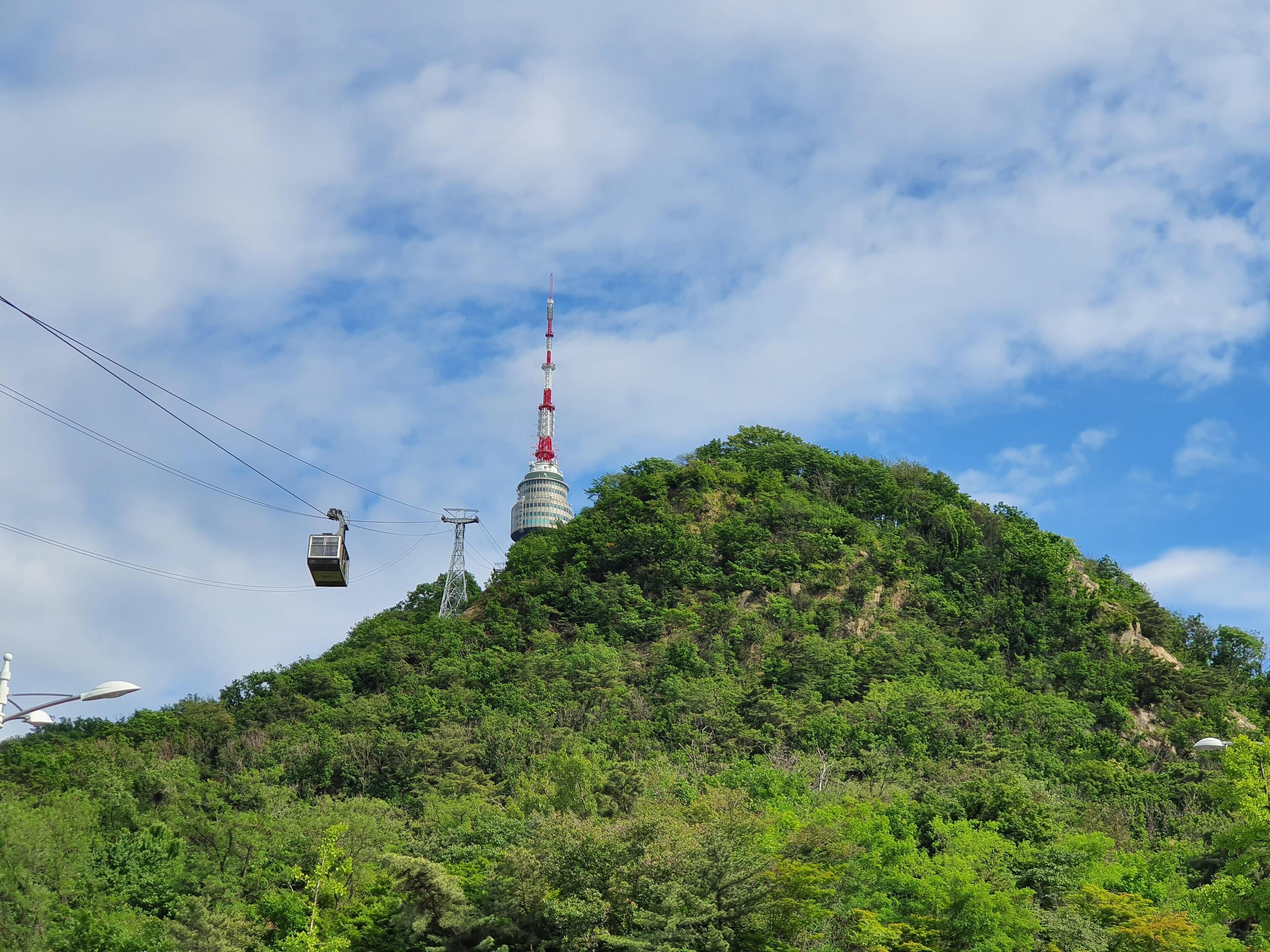 Un téléphérique traverse une colline verdoyante, avec une tour imposante qui se détache sur l’horizon.