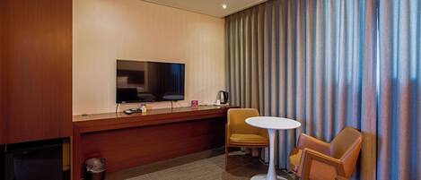 Una habitación de hotel con televisión, escritorio, silla, una mesita y cortinas.