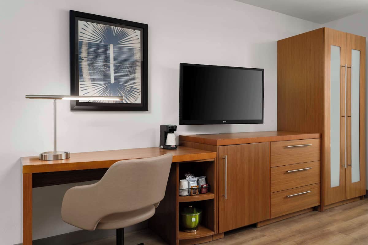 A modern room with a wooden desk, a chair, a flat-screen TV, a coffee maker, and a cabinet with drawers.