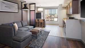A modern apartment with a grey sofa, a small dining area, a kitchen with a TV, and a large window offering a city view.