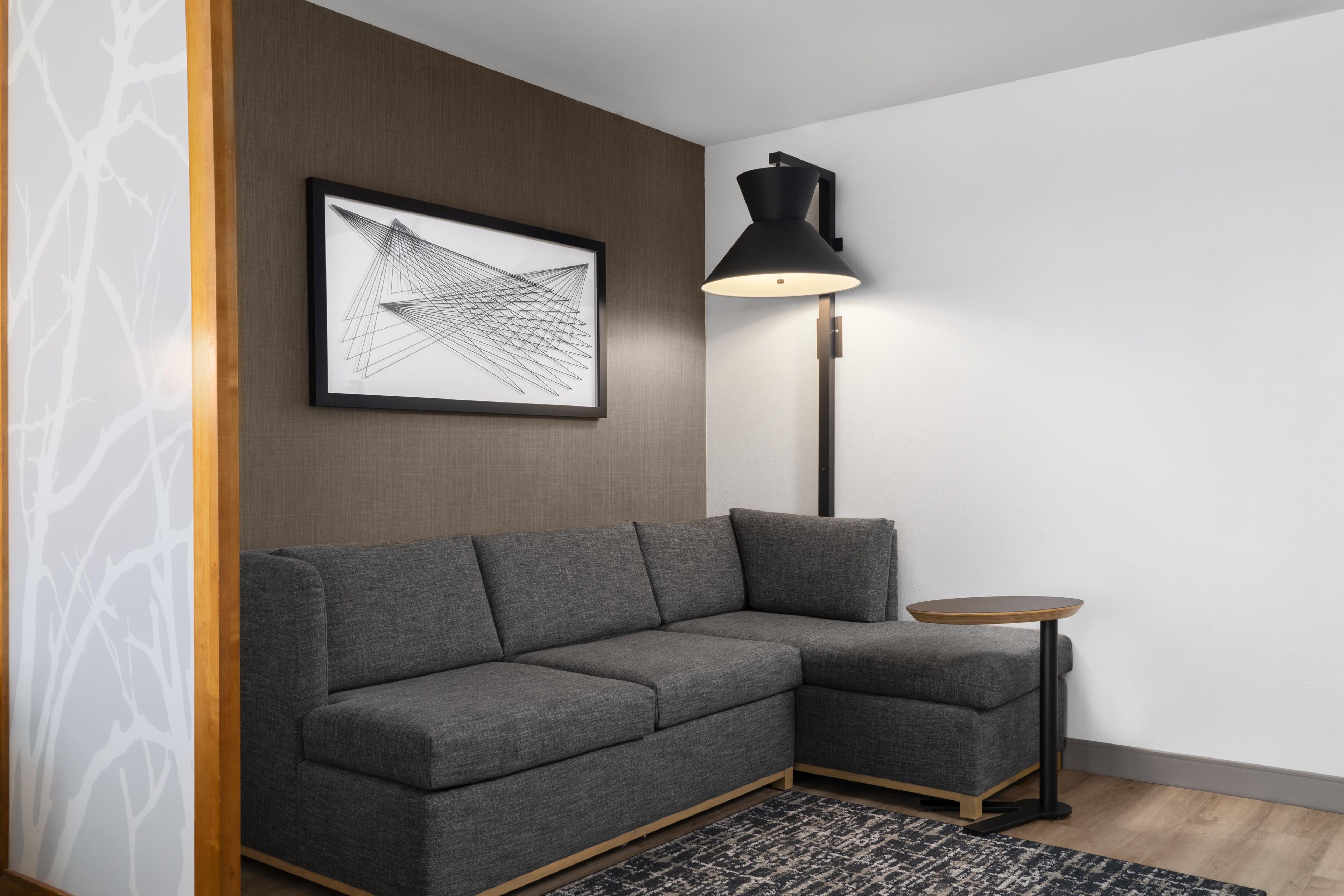 A modern living room with a grey sectional sofa, a small round side table, a floor lamp, and a framed abstract artwork on the wall.