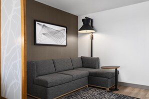 Egy modern nappali, melyben egy szürke, moduláris kanapé, egy kis, kerek dohányasztal, egy állólámpa és egy keretezett, absztrakt műalkotás található a falon.
