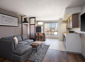 Un appartement moderne avec un canapé gris, une petite table  à manger, un coin cuisine et une grande fenêtre donnant sur la ville.