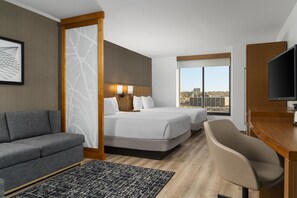 Habitación de hotel con sofá, cama con dos almohadas, escritorio y televisor, y vista a la ciudad a través de una ventana.