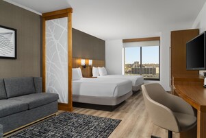 Une chambre d’hôtel avec un canapé, un lit avec deux oreillers, un bureau avec une télévision et une vue sur la ville par la fenêtre.