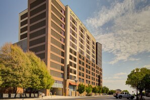 Un hotel de varios pisos con un letrero llamativo de "Hyatt Place", un estacionamiento subterráneo y vista a la calle con árboles y un camión estacionado.