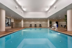 Une grande piscine intérieure avec des bords carrelés, entourée de chaises longues et d’un espace détente comprenant des tables et des chaises.
