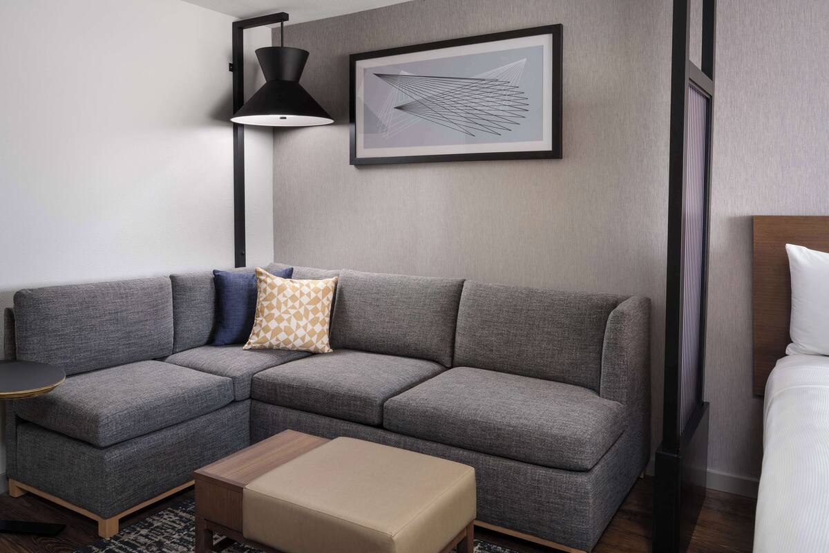 A modern hotel room with a grey sectional sofa, a small wooden side table, a black floor lamp, and a framed abstract artwork on the wall.