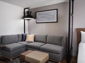 Un salon moderne avec un canapé d’angle, une petite table en bois, un lampadaire et une œuvre d’art abstraite encadrée accrochée au mur.