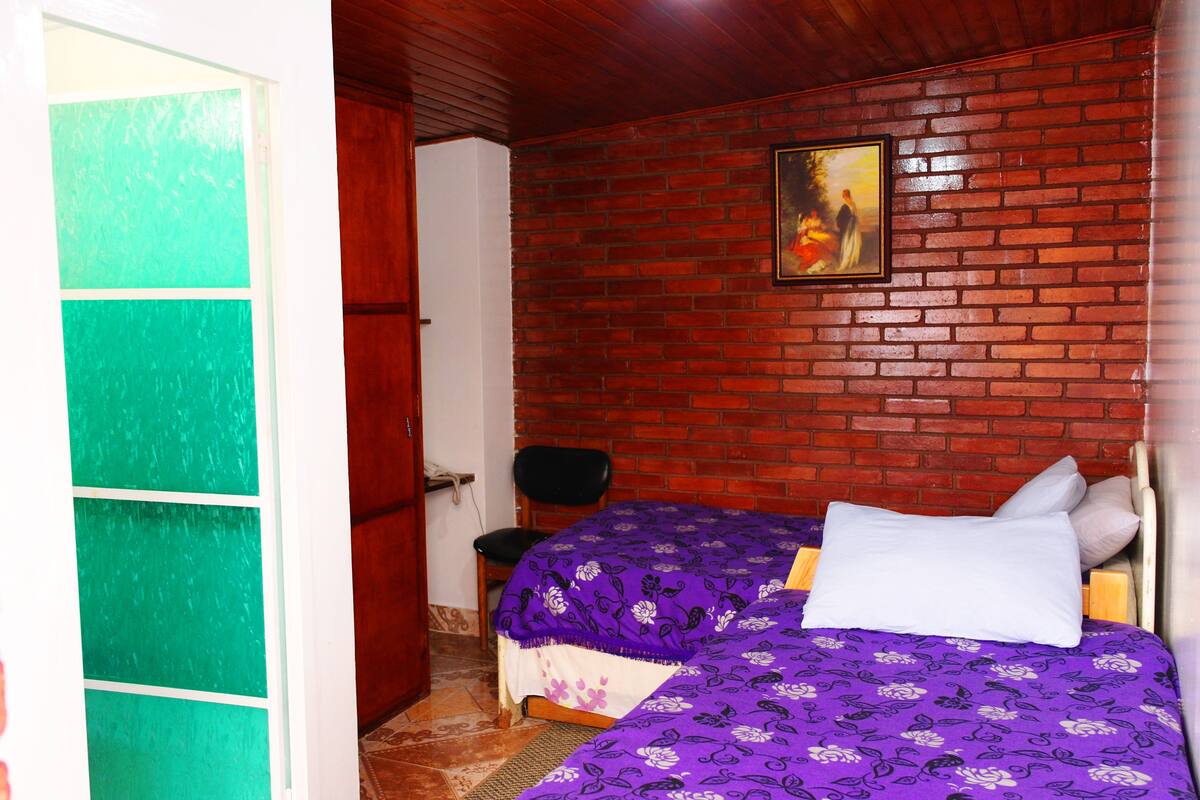 A room with a brick wall, a bed with a purple floral bedspread, a white pillow, and a framed picture on the wall.
