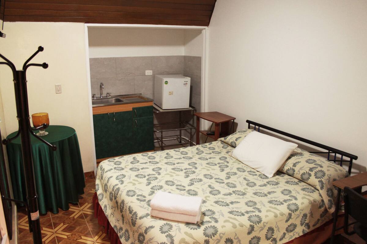 A bedroom with a bed, a nightstand, a table with a lamp, and a kitchenette in the background.