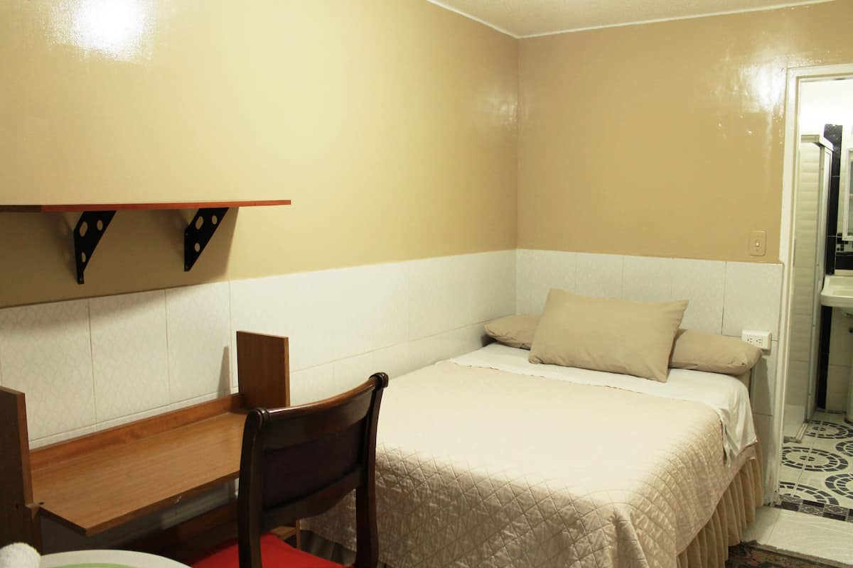 A small, simple room with a bed, a desk, and a chair. There is a shelf above the desk and a door to the right.