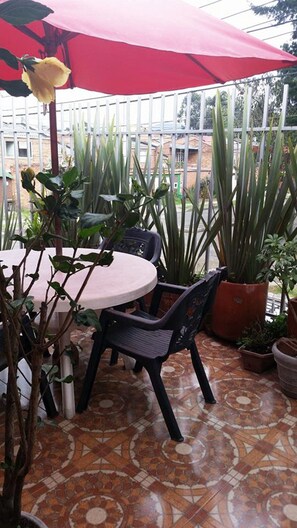 Een terras buiten met een ronde tafel, twee stoelen, een rode parasol en diverse potplanten.