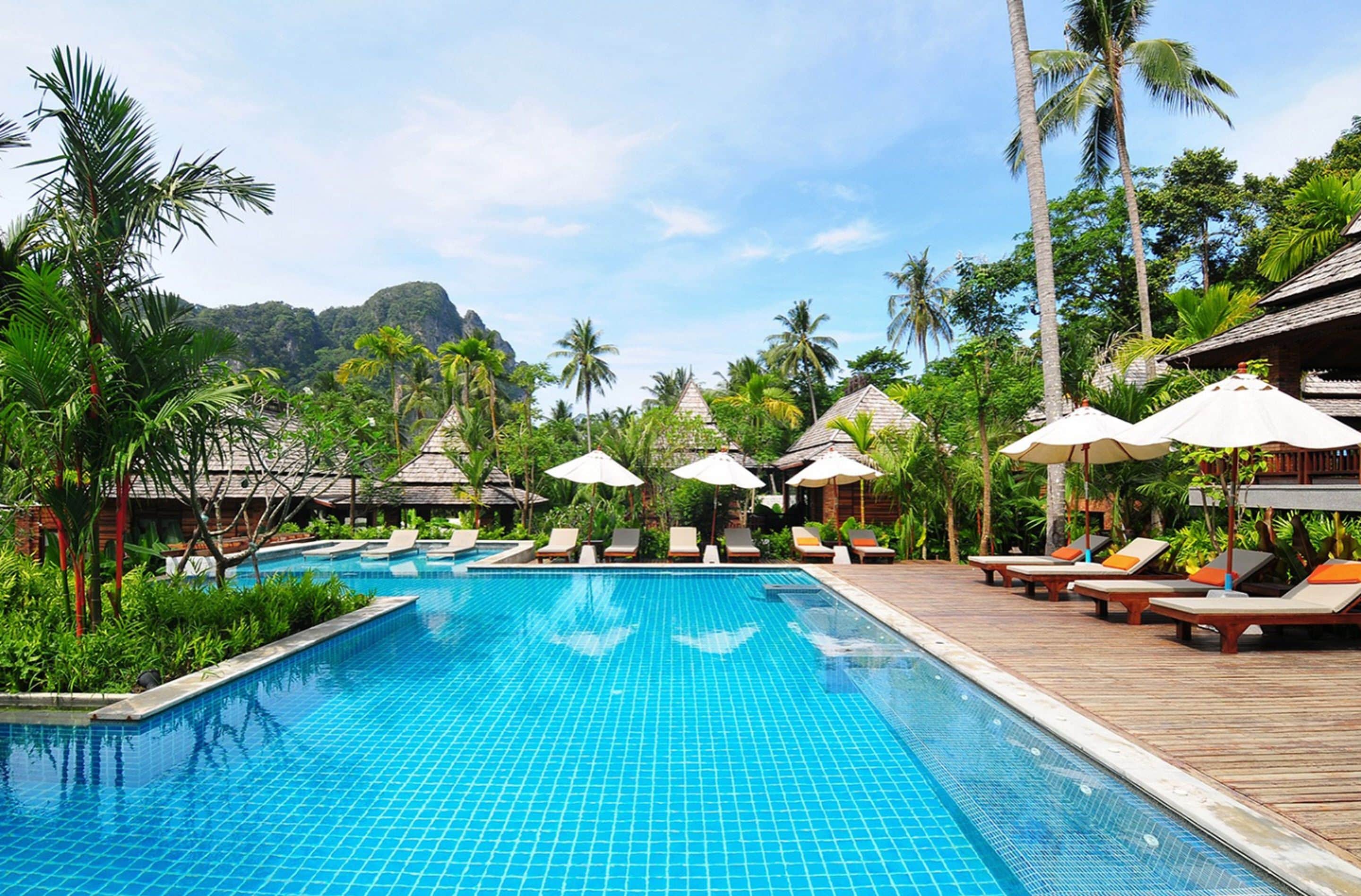 A swimming pool with lounge chairs and umbrellas, surrounded by palm trees and a mountain in the background.