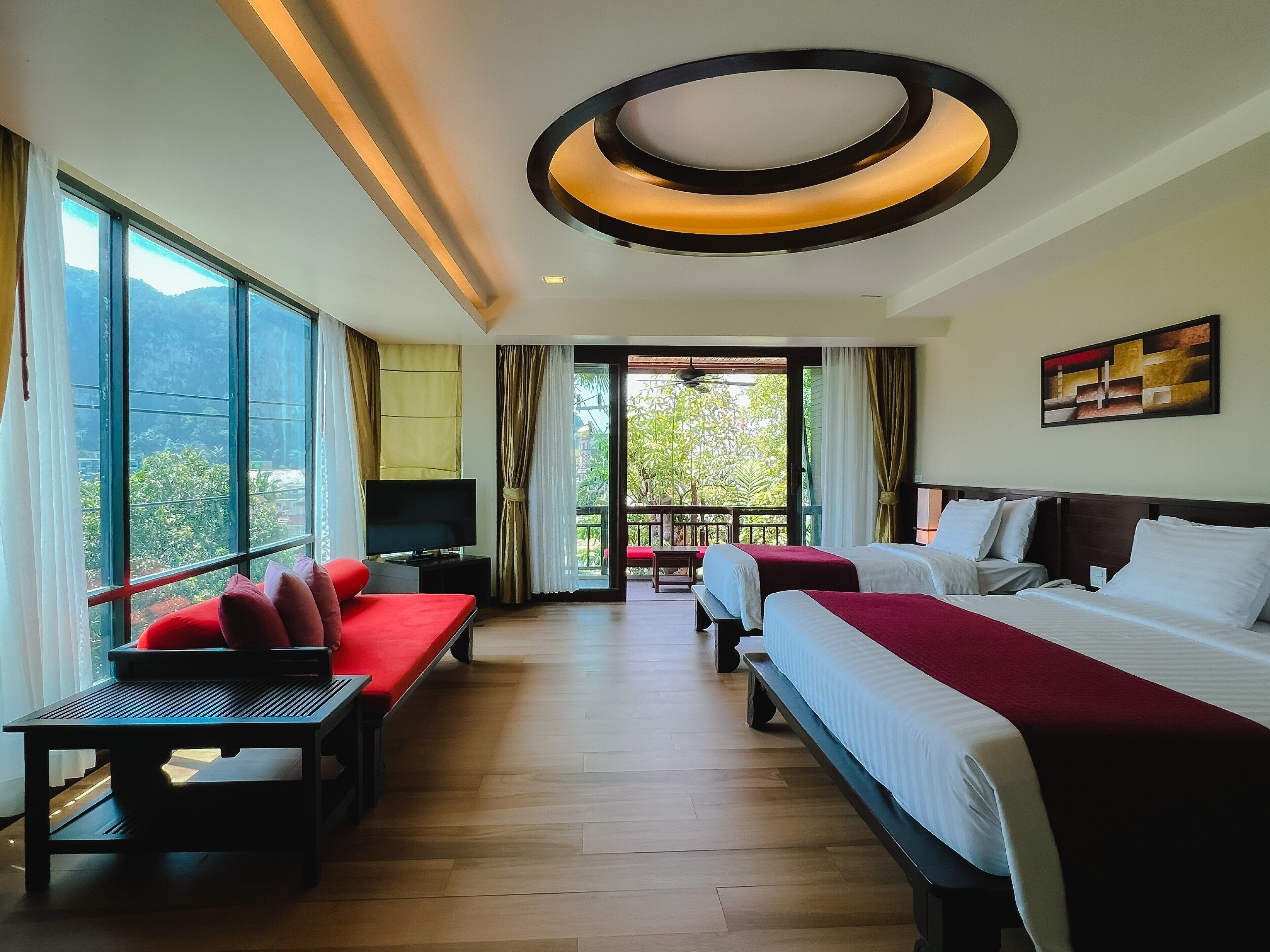 A hotel room with two beds, a TV, a desk, and a balcony with a view of greenery.