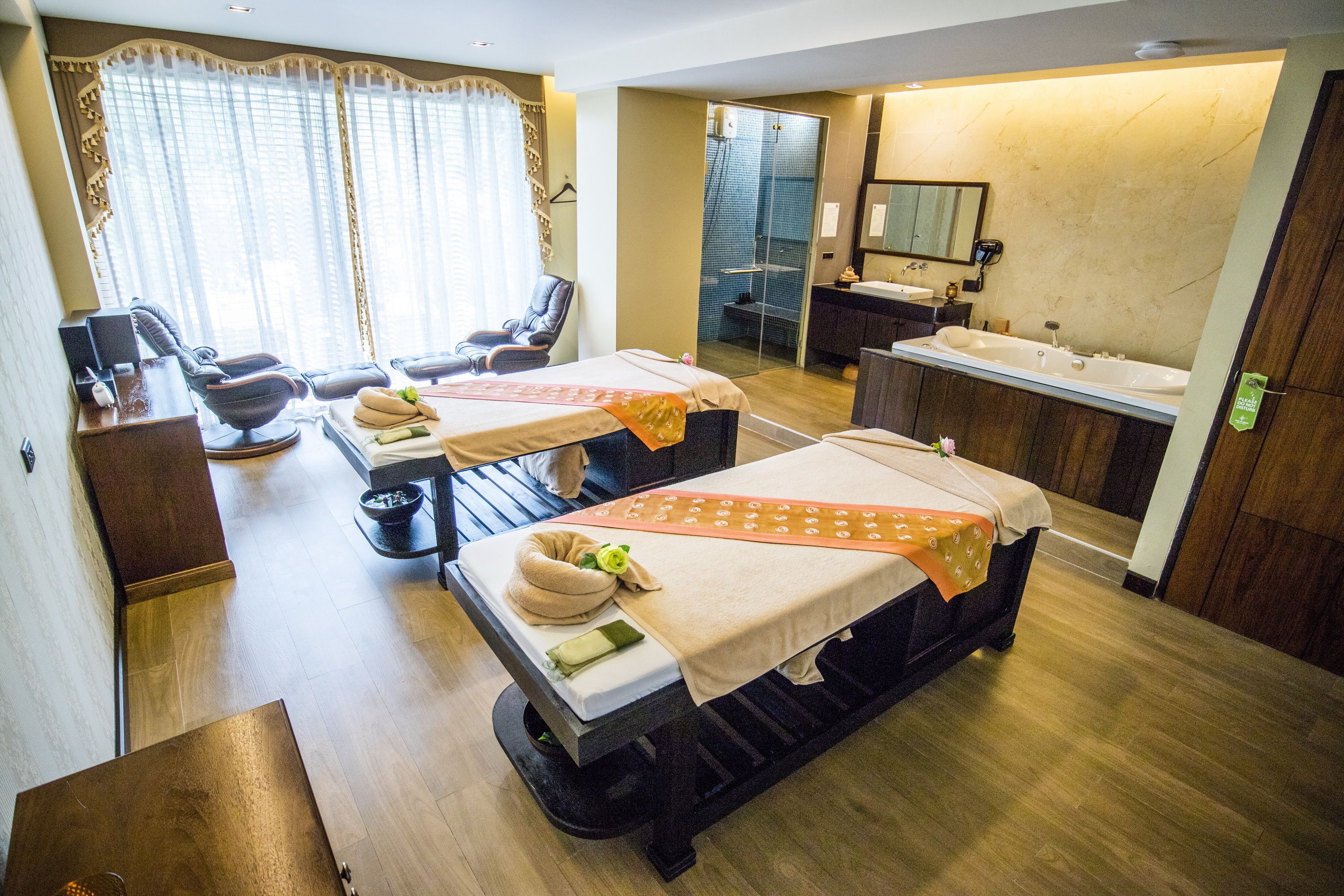 Une chambre d’hôtel équipée de deux tables de massage, d’un fauteuil de massage séparé, d’un espace douche et d’une salle de bain avec un grand miroir et un lavabo.