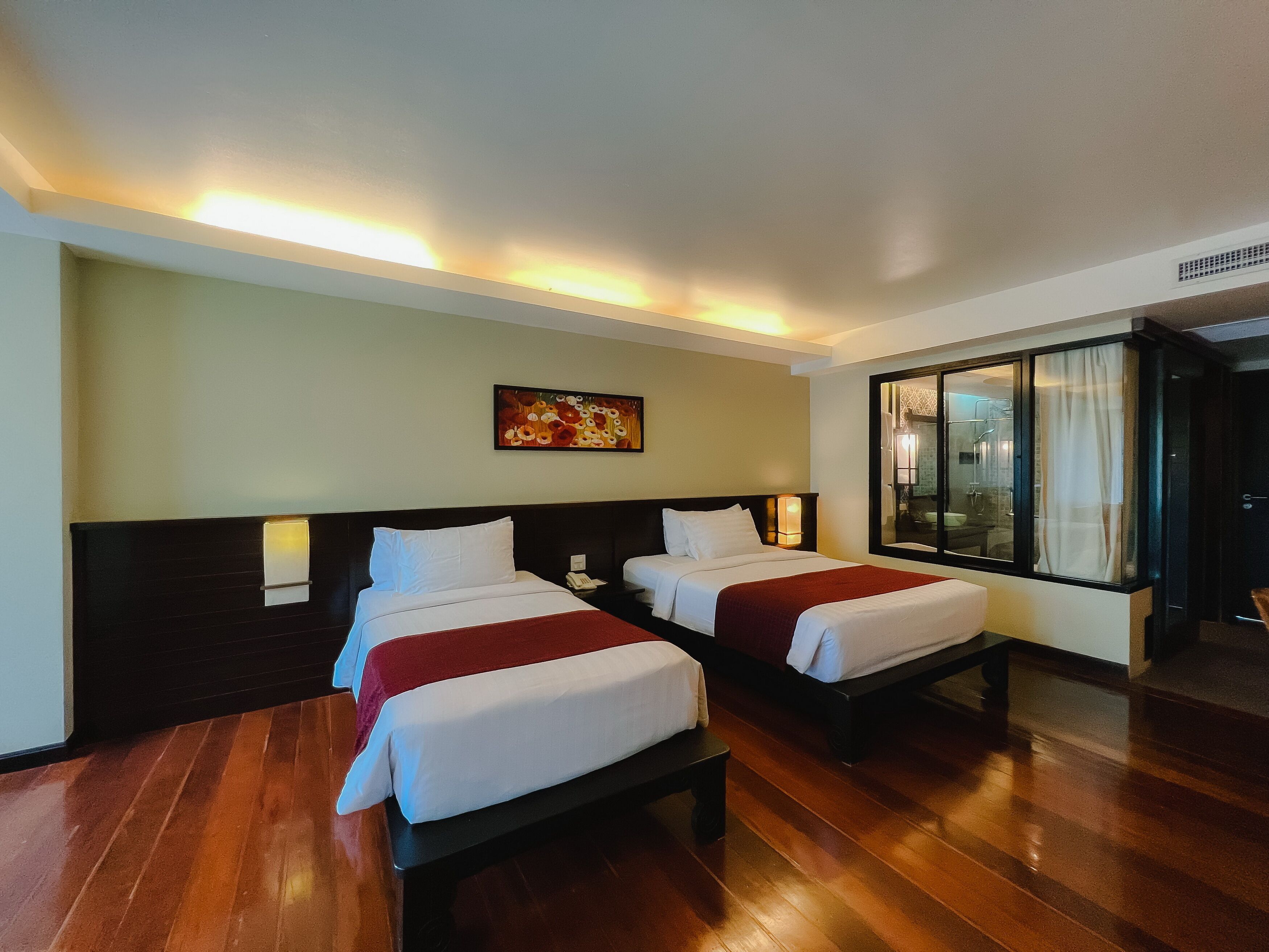 A hotel room with two beds, a painting on the wall, and a window with a view of the bathroom.