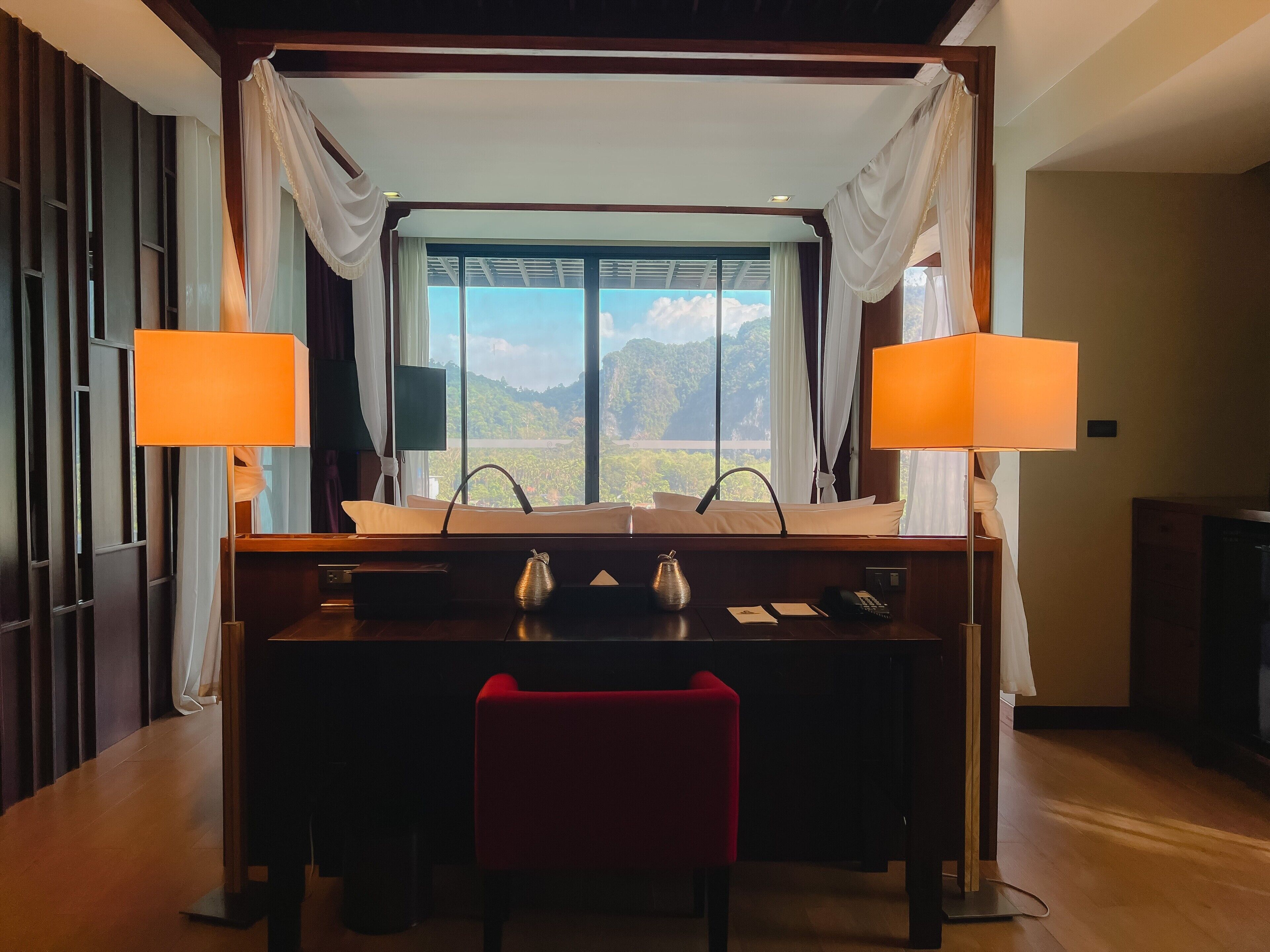Quarto de hotel com uma janela grande que oferece vista para as montanhas, uma mesa de jantar de madeira com duas lâmpadas, uma cadeira vermelha e um minibar.