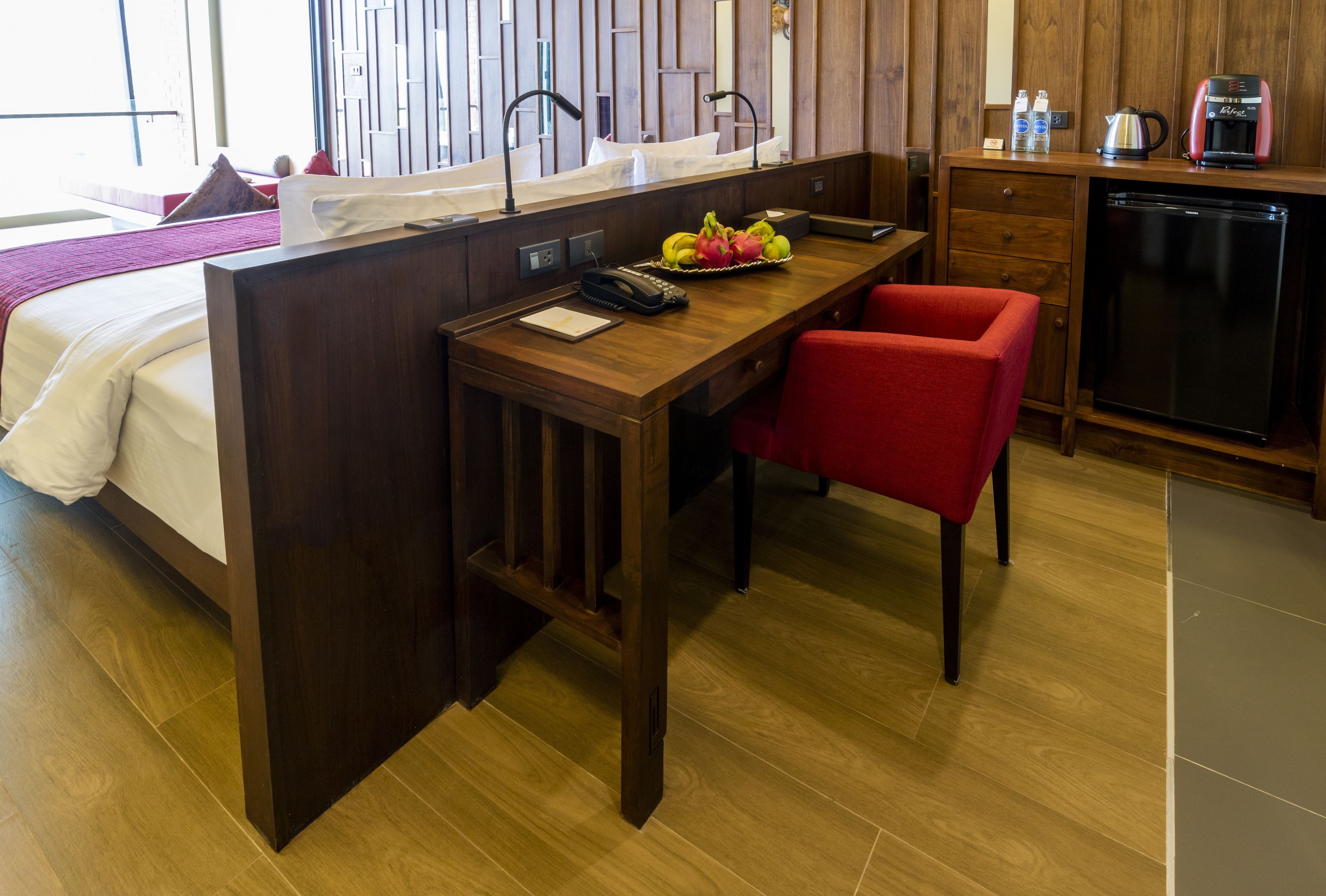 A hotel room with a wooden desk, a red chair, a small kitchen area with a coffee maker, and a bed with white linens.