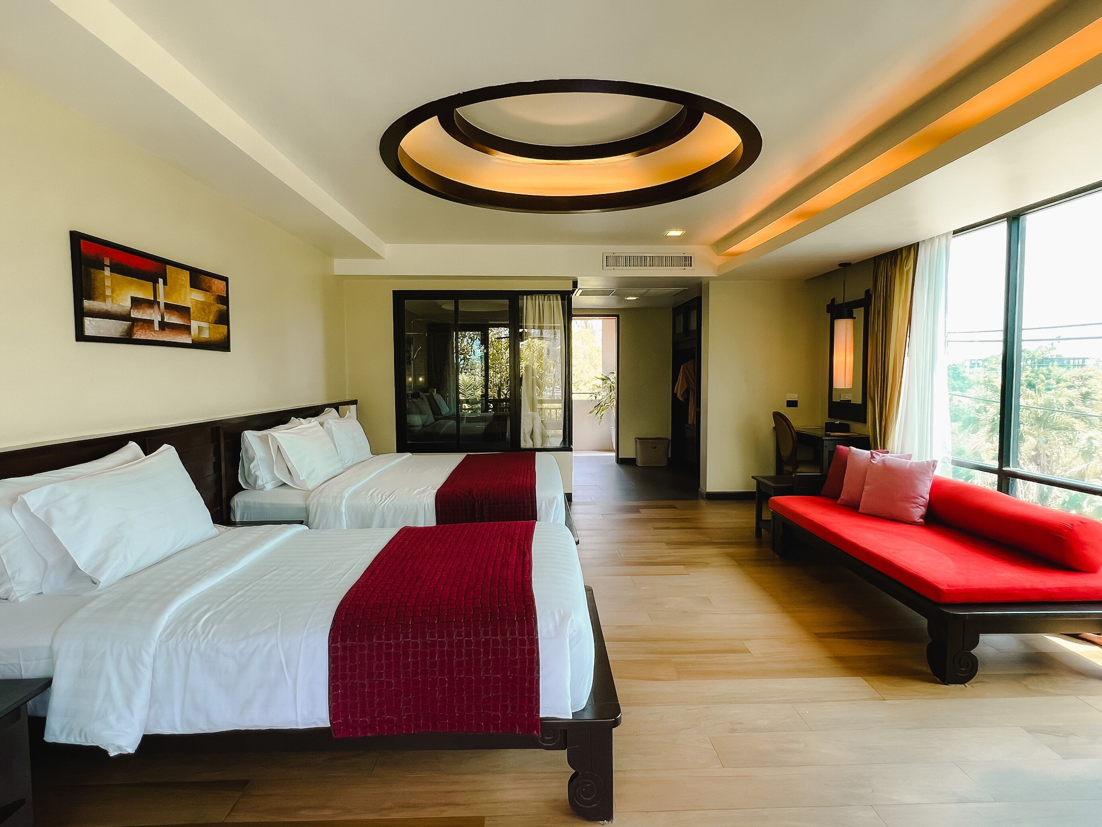 A hotel room with two beds, a red sofa, a large window, and a ceiling light fixture.