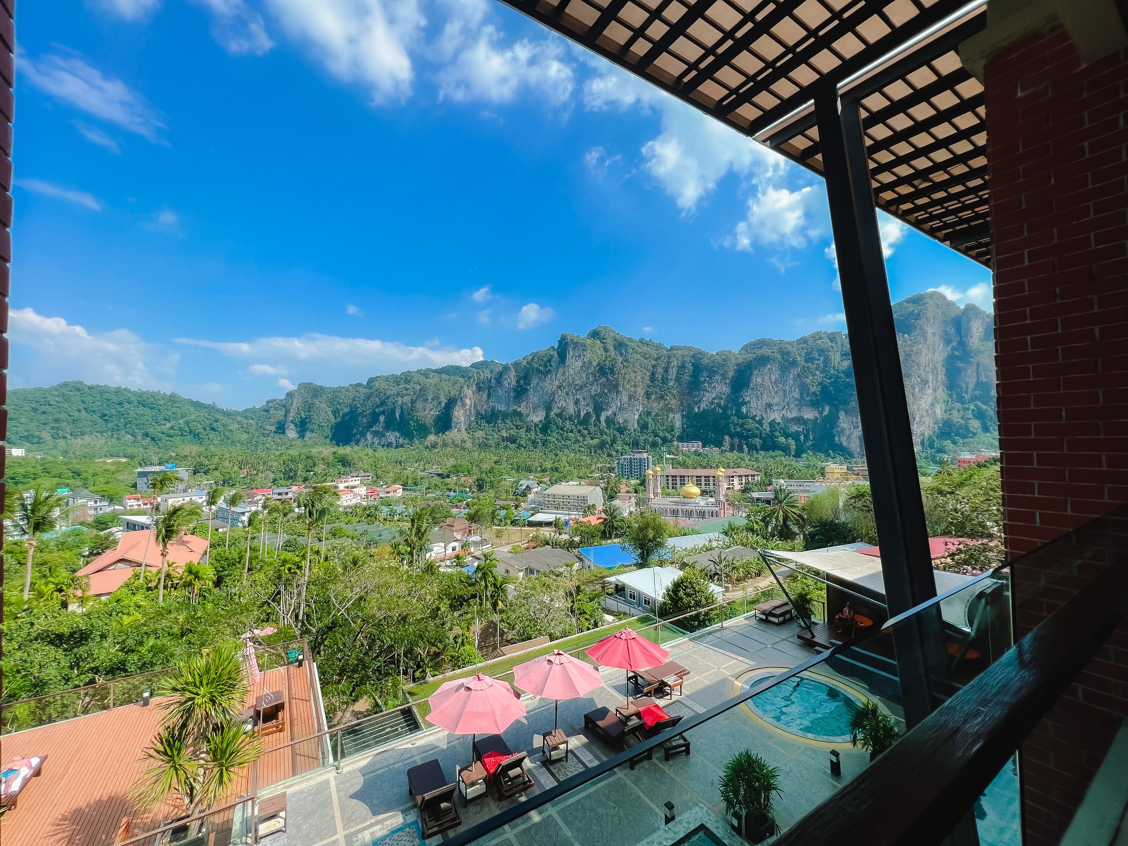 A balcony with a pool, lounge chairs, and umbrellas, overlooking a town with mountains in the background.