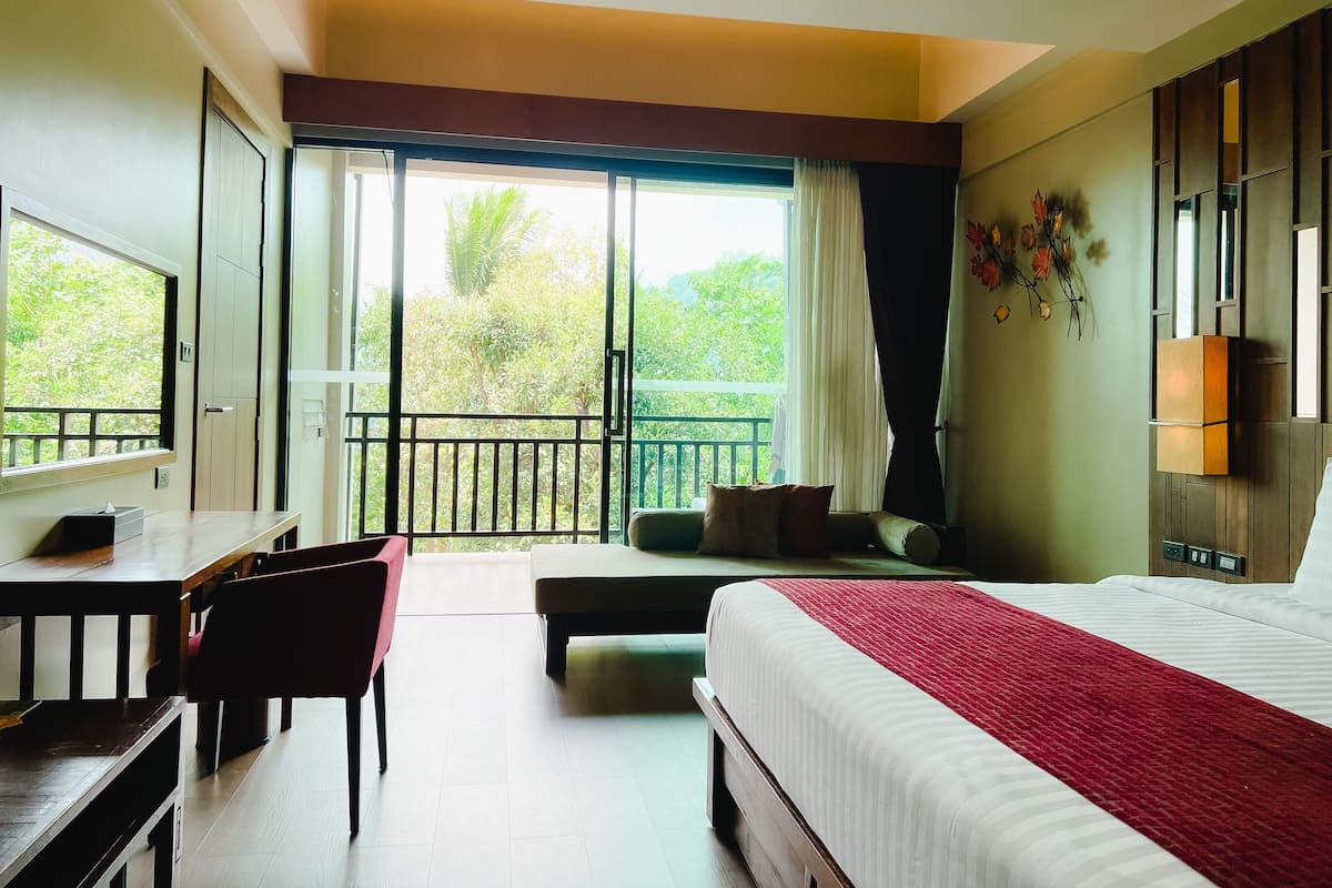 A hotel room with a bed, a desk, a chair, a balcony with a view of greenery, and a window with curtains.