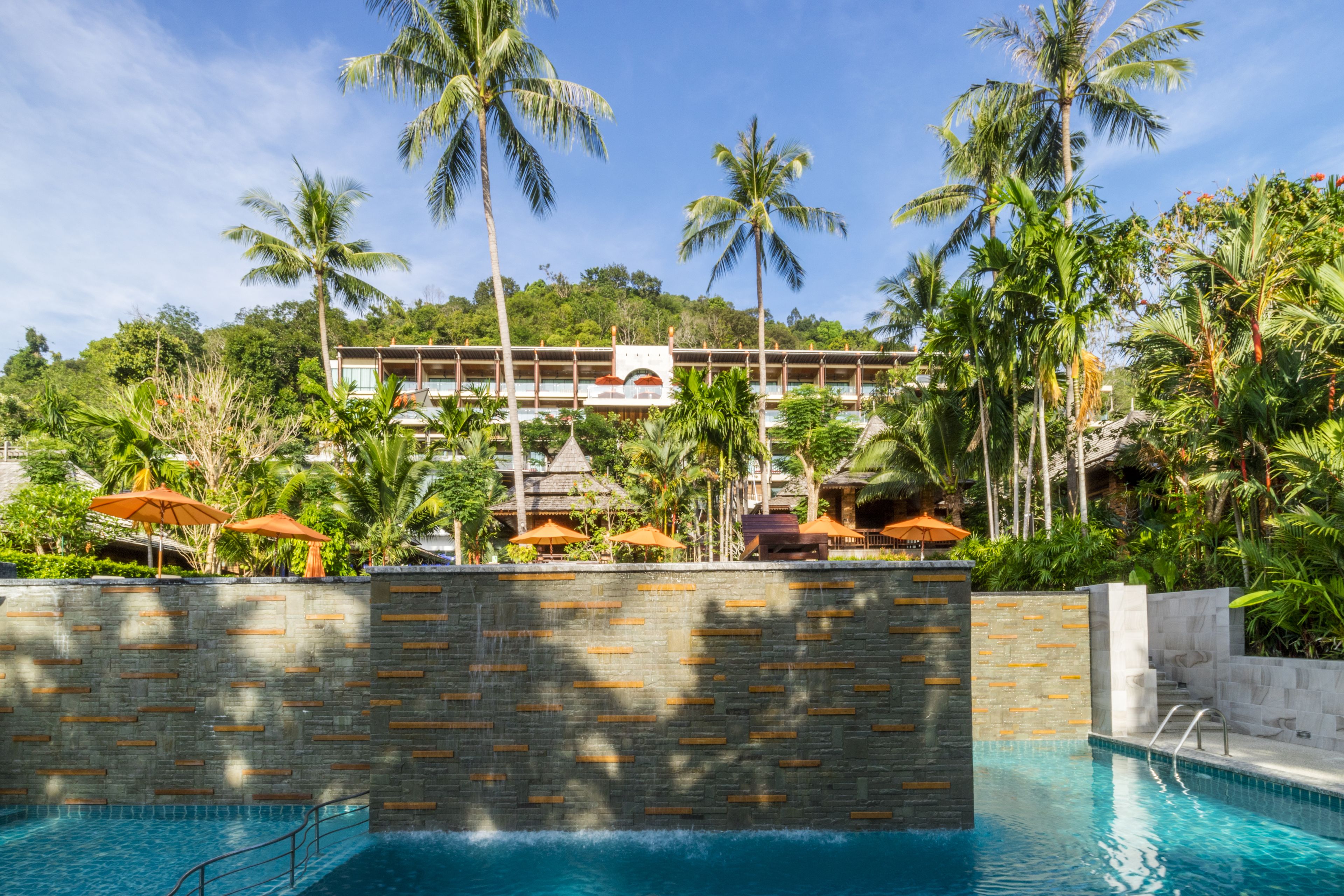 A pool area with a stone wall, palm trees, and a building in the background.