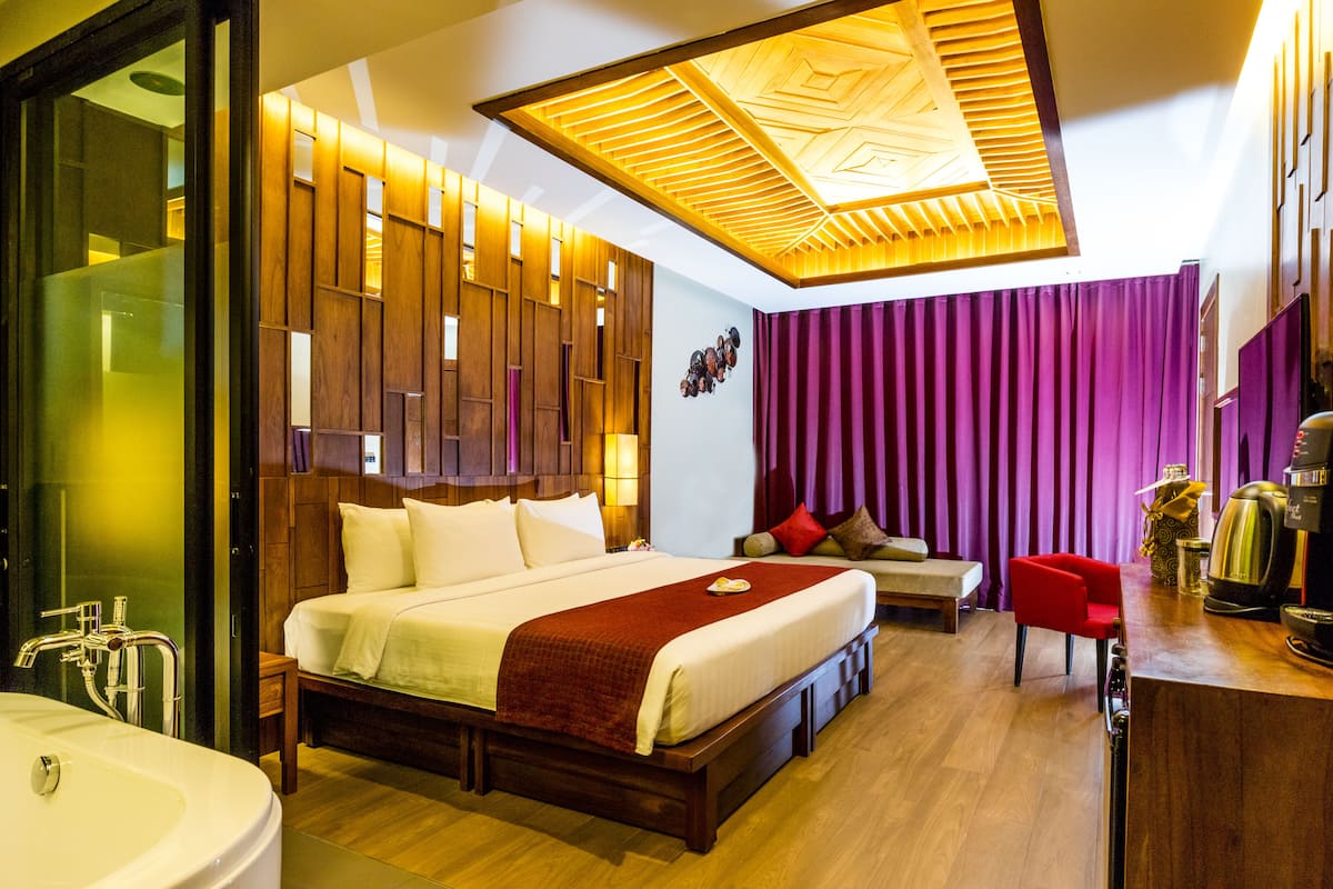 A modern hotel room with a large bed, a wooden headboard, a red chair, a coffee maker, and a large window with curtains.