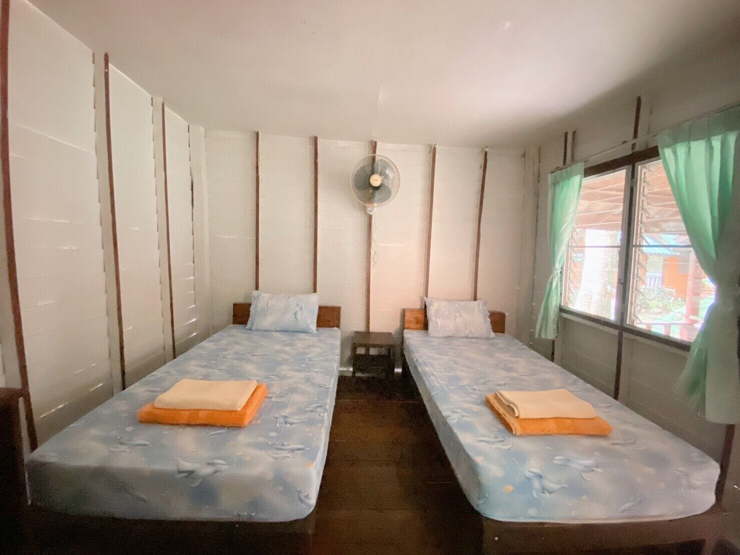 A room with two single beds, a small table, a fan, and a window with curtains.